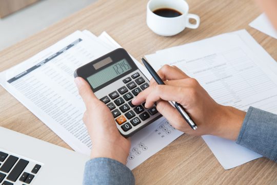 Unrecognizable businessman using calculator to find out income and outcome. He counting company finances. Male hands with pen pushing button at desk. Investment or paperwork concept